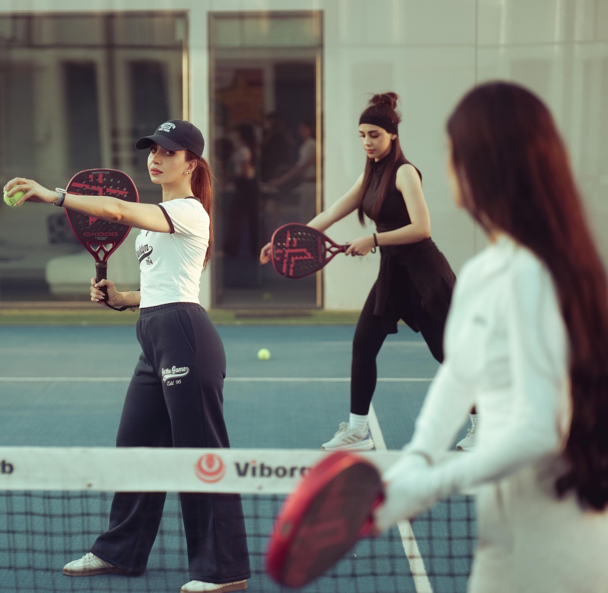 Three people playing padel on an outdoor court, holding bats and preparing to hit a ball over the net during a game.