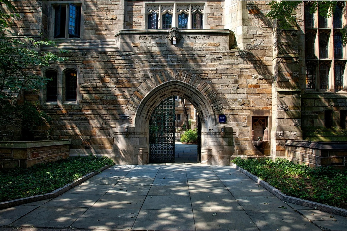 Stone archway entrance to a historic building with detailed masonry, iron gate, and greenery lining the walkway.