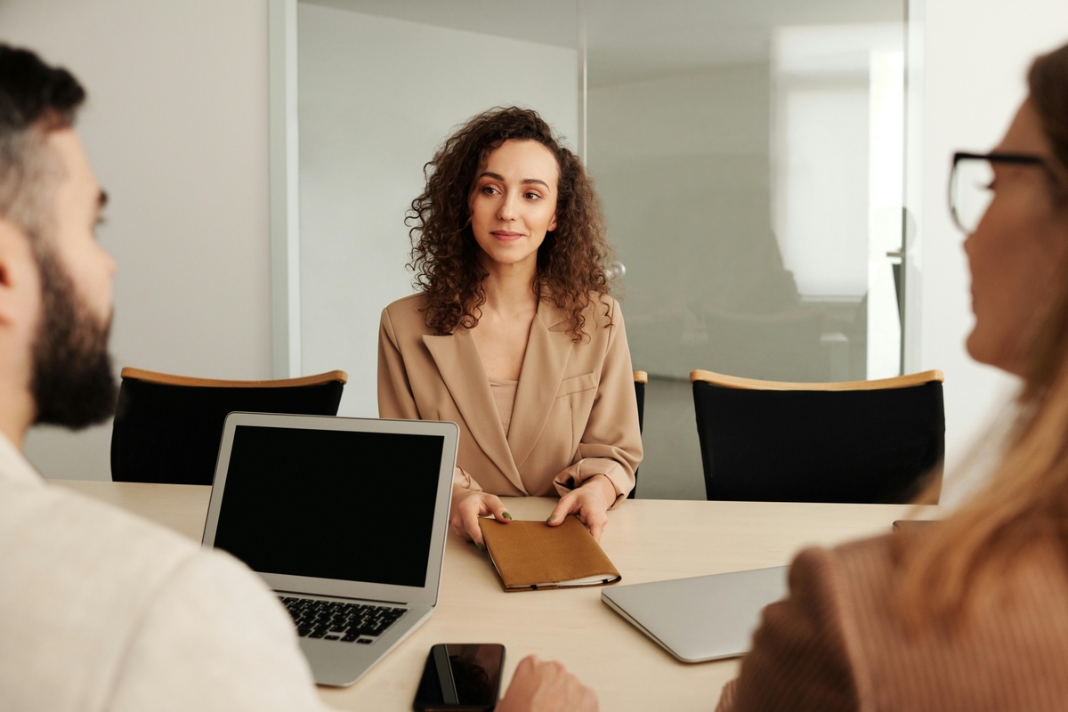 Two people sit at a conference table with laptops while another person sits across from them holding a notebook in a modern meeting room.