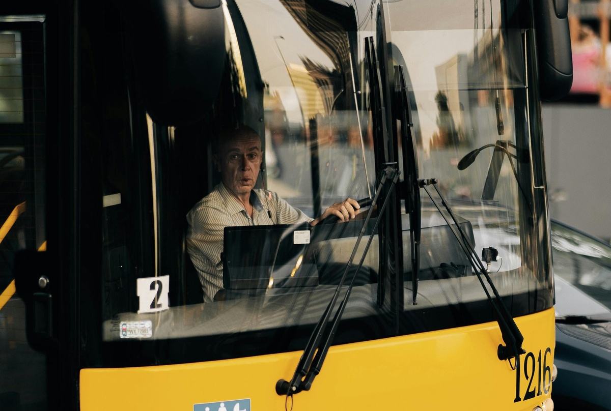 Person seated in the driver’s seat of a yellow city bus, holding the steering wheel, viewed through the front window with street reflections visible on the glass.