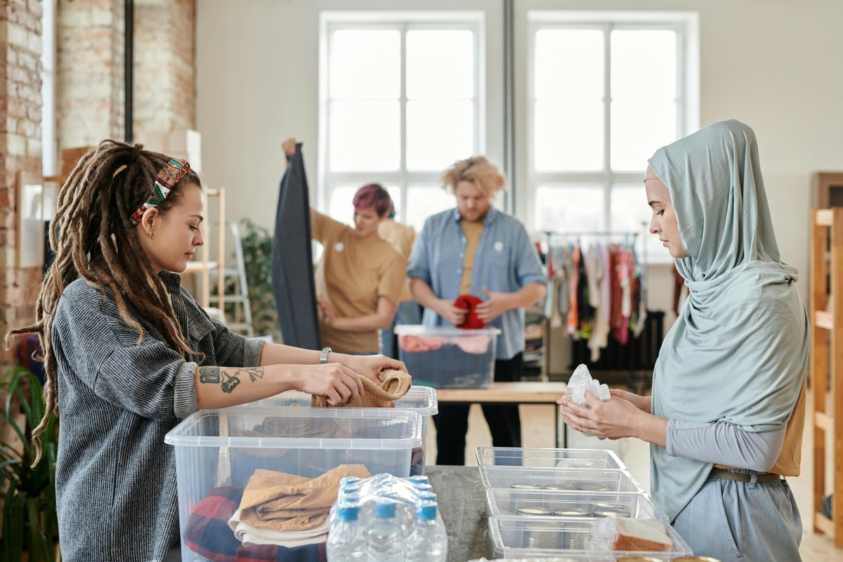 Group of people sorting and folding clothing at tables in a bright indoor space, with clear plastic storage boxes, water bottles, and racks of clothes visible in the background.