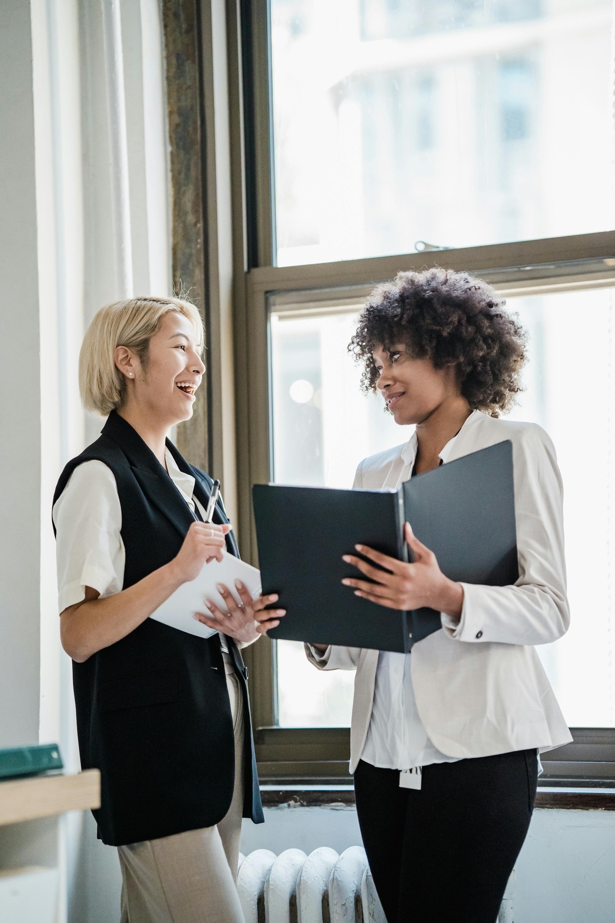 Two people standing in a bright office space, holding open folders and talking together near a window, with natural light illuminating the scene.