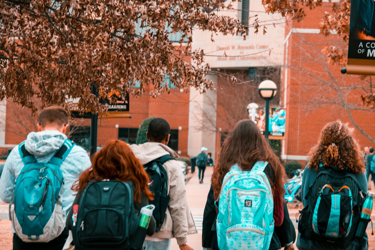 The image captures a group of individuals walking outdoors on what appears to be a school or university campus. The scene is set along a paved pathway bordered by buildings made of red brick. 