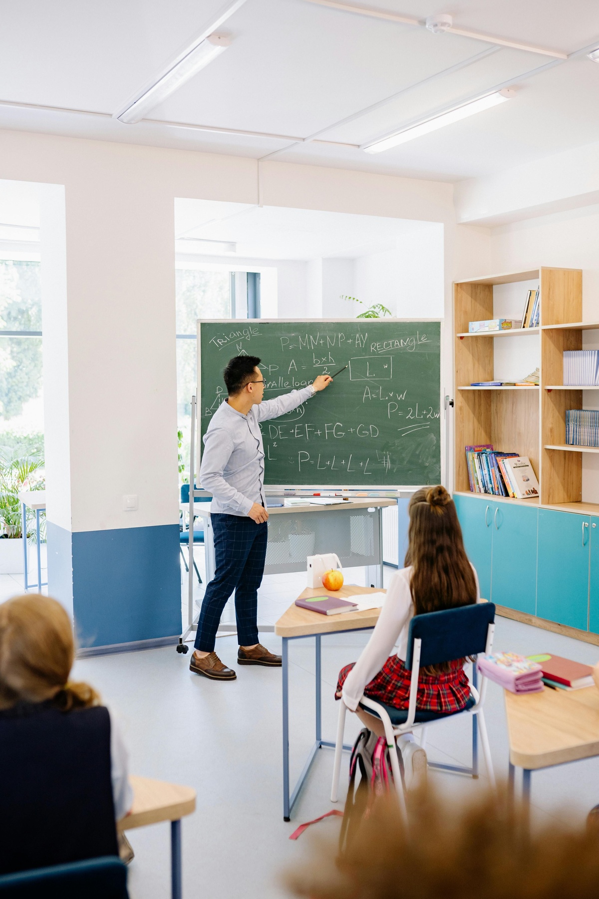 Teacher standing beside a chalkboard filled with maths equations, pointing to the board while a student sits at a desk facing forward in a classroom setting.