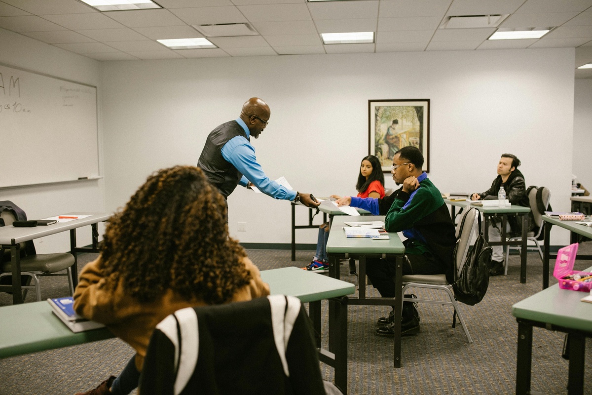 A person at the front of a classroom hands a paper to a student while several others sit at desks in a bright, organized room.