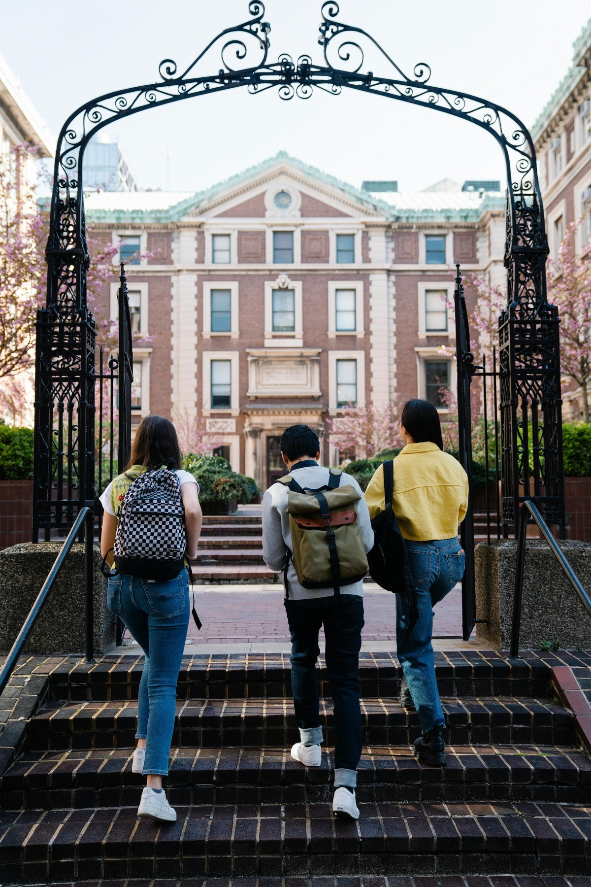 Three people with backpacks walk up steps through an ornate iron gate toward a large brick academic building with arched windows.