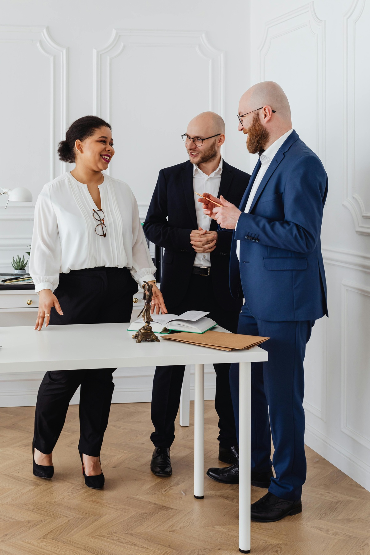 Three people standing around a table in a bright office, discussing together, with notebooks, a desk ornament, and documents on the table.