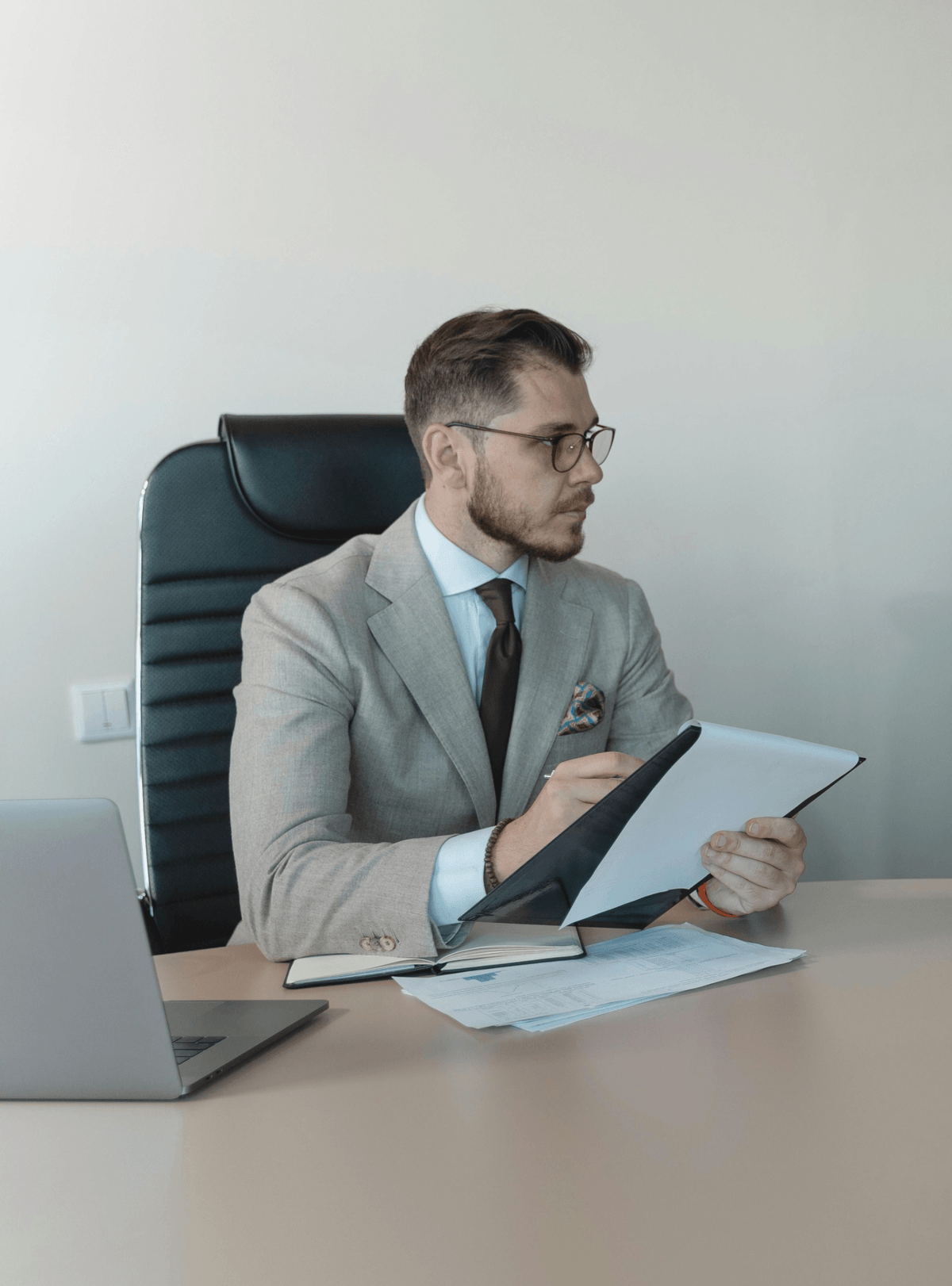 The image shows an indoor office setting with a person seated at a desk. The individual is wearing a light-colored suit jacket over a dress shirt and tie. The person is holding a clipboard or notepad in one hand and appears to be writing or reviewing information with a pen in the other hand.