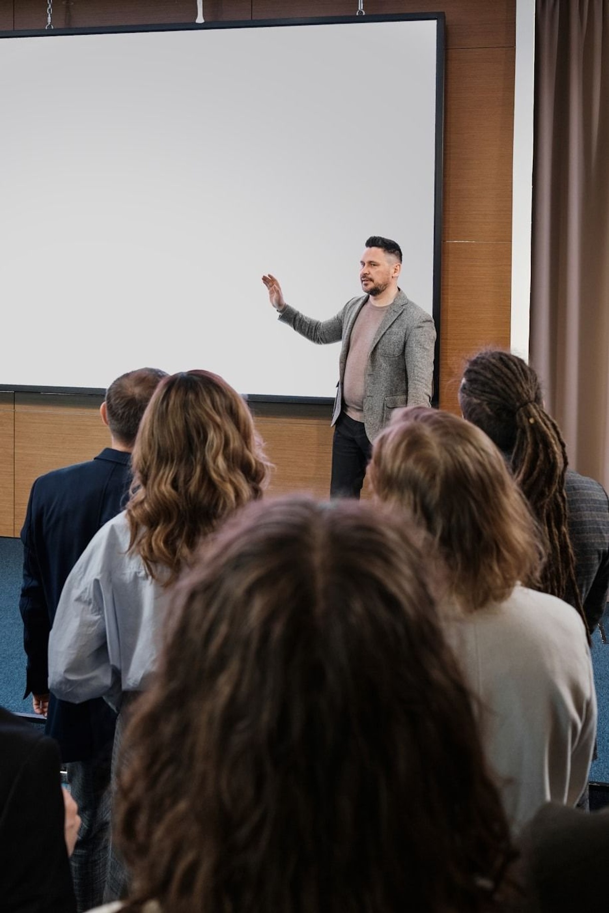 A person stands at the front of a room giving a presentation, gesturing toward a large screen, while a group of people face forward listening.