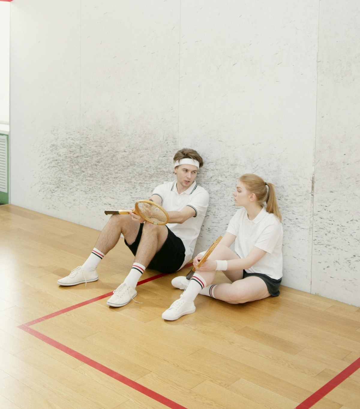 Two people sitting on the floor of an indoor sports court, holding rackets and resting against a wall, with wooden flooring and court markings visible.