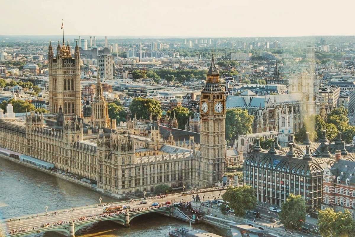 A wide aerial view of the Palace of Westminster beside the River Thames, including the clock tower commonly known as Big Ben, with the cityscape of London extending into the distance.
