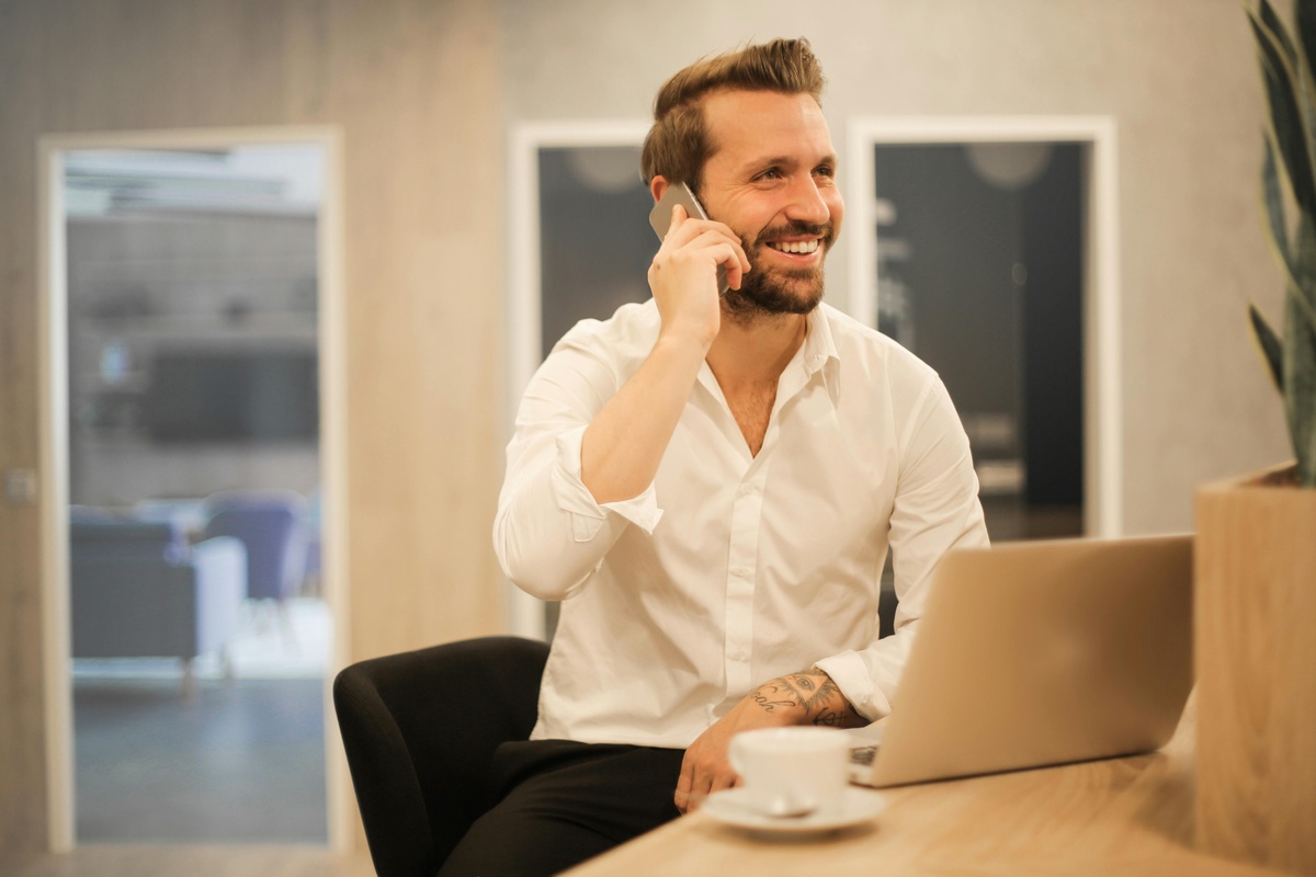 A person in a white shirt sits at a desk with a laptop and a cup of coffee, holding a phone to their ear in a modern office setting.