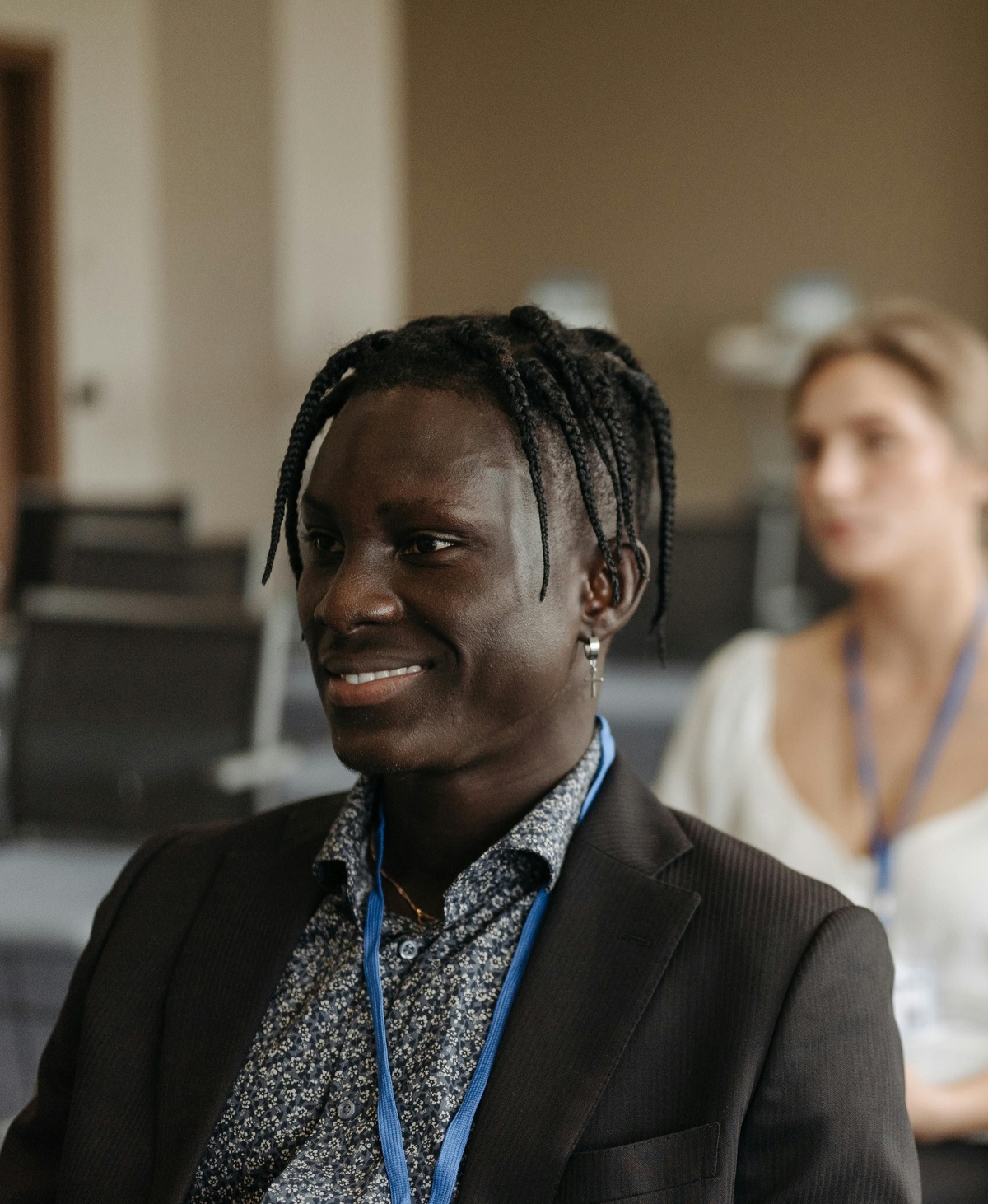 Person seated in a lecture or training room wearing a lanyard, looking forward while another attendee sits slightly out of focus in the background among rows of seats.