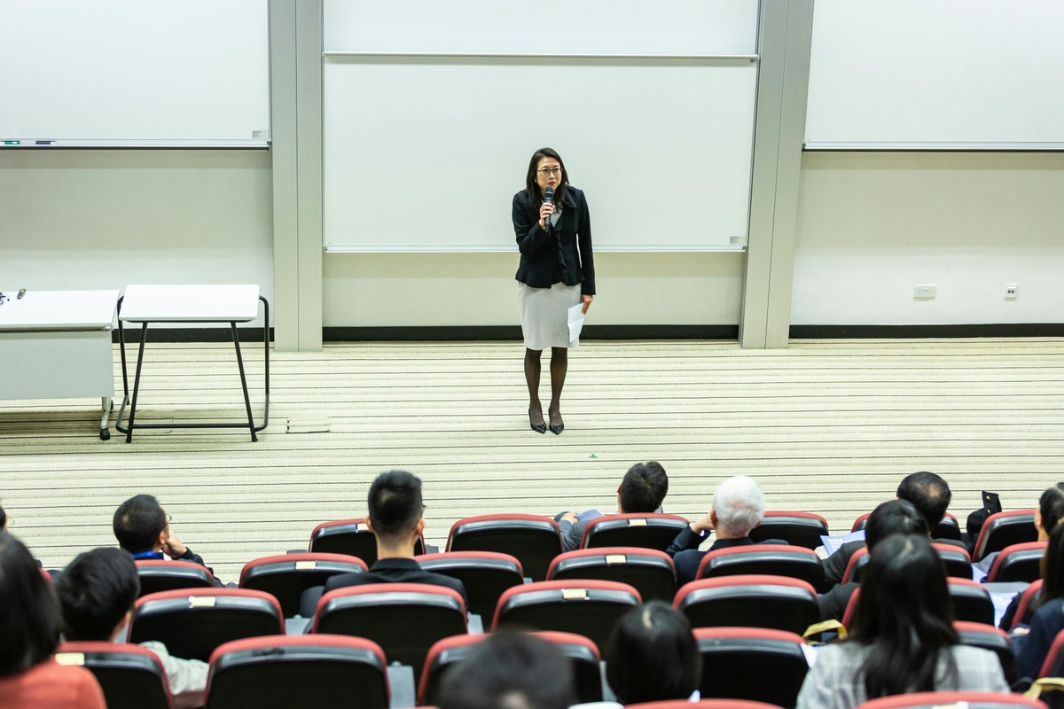A person stands at the front of a lecture hall speaking into a microphone, facing an audience seated in tiered rows.