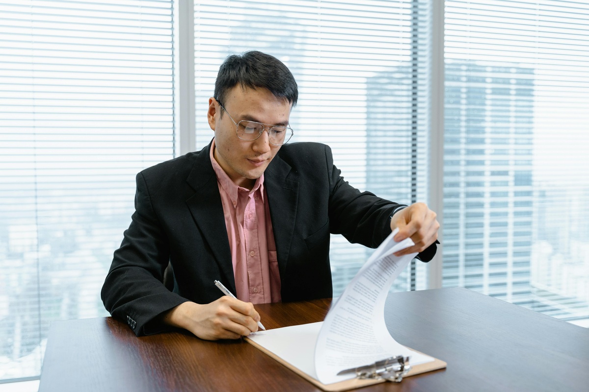 A person in business attire sits at a desk in a bright office, holding a document in one hand and writing on a clipboard with the other. Large windows behind the person show a cityscape.