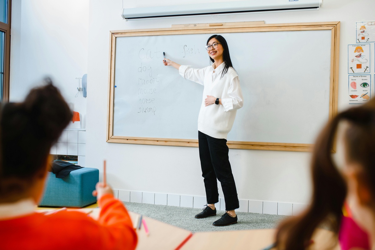 An individual stands at a whiteboard holding a marker and pointing to written text, while several students sit at desks in the foreground facing the board.