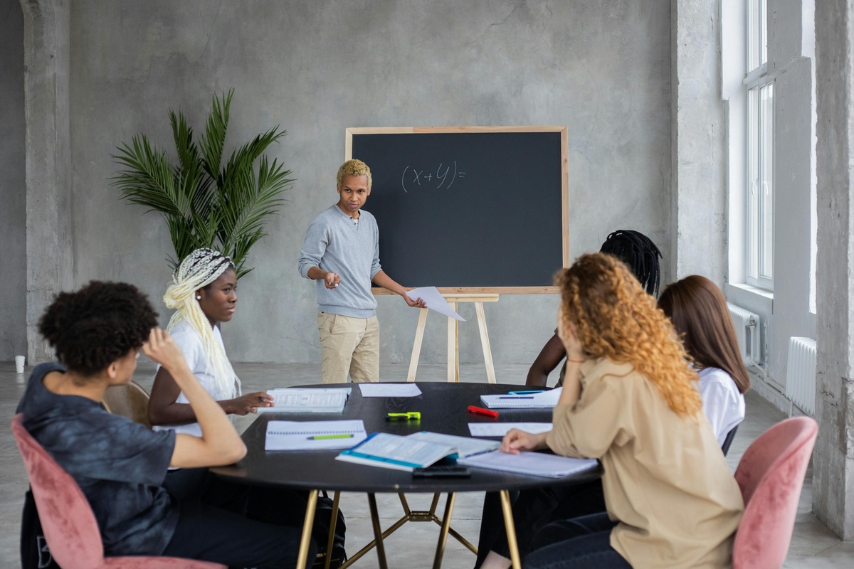A person stands at a chalkboard explaining an equation to a group seated around a table covered with notebooks and papers in a modern classroom.