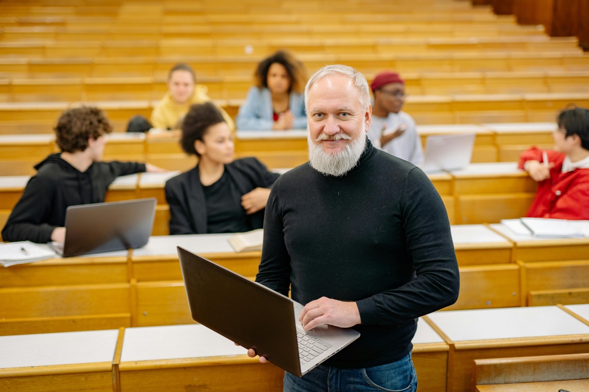 A person stands in a lecture hall holding a laptop while several others sit in the background with their own laptops, talking among themselves.