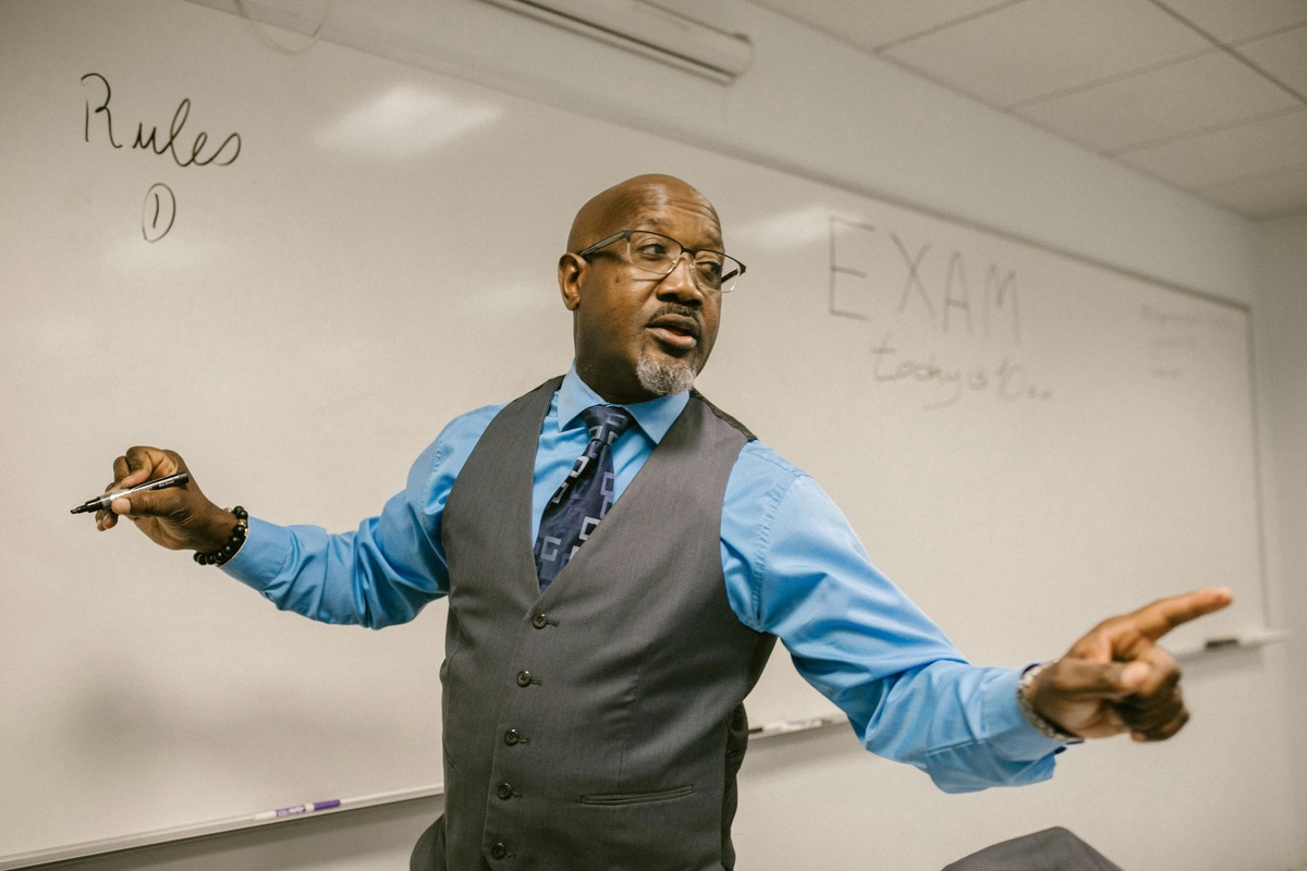 A person stands in front of a whiteboard holding markers, gesturing with both hands as if teaching or presenting, with the word “EXAM” written on the board behind them.