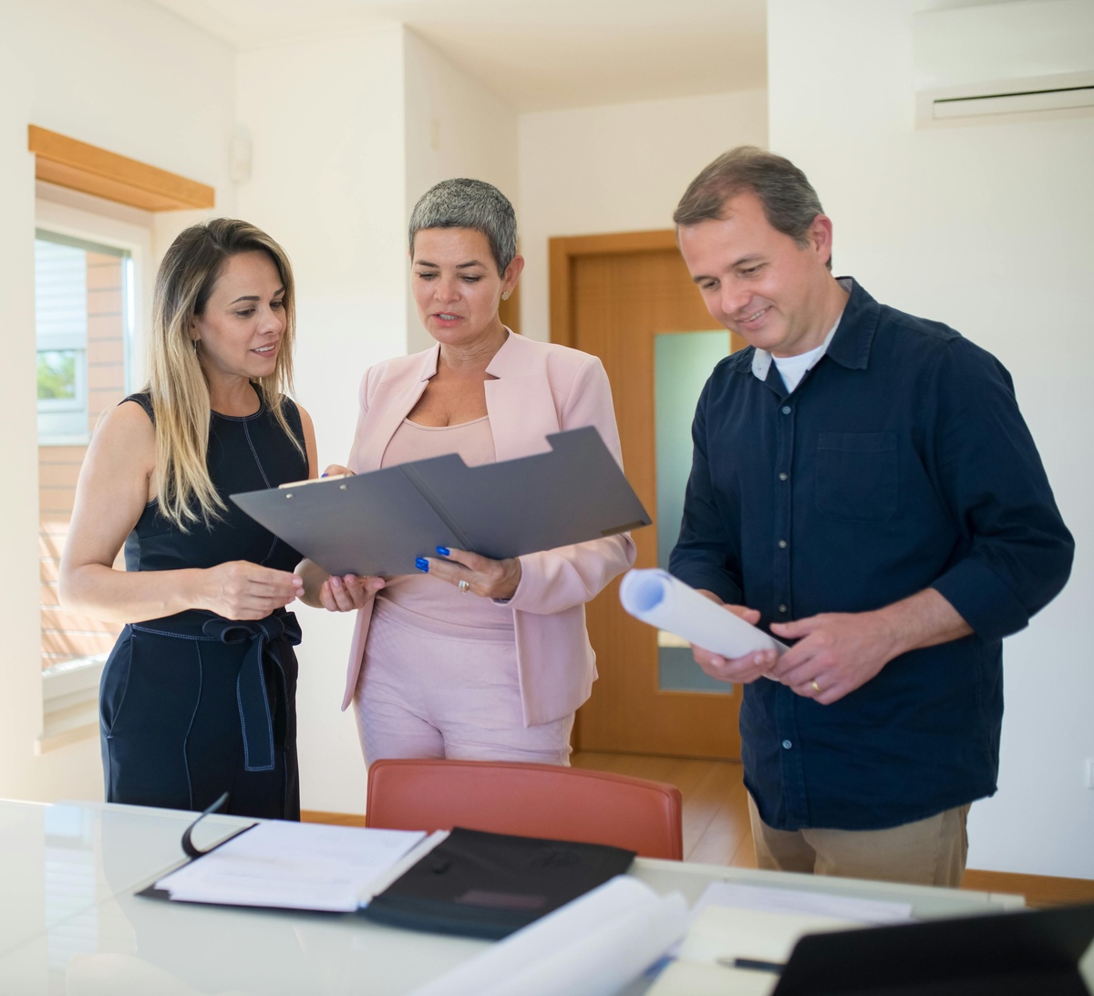 Three people standing around a table in a bright office, reviewing documents together, with folders and papers laid out on the table.