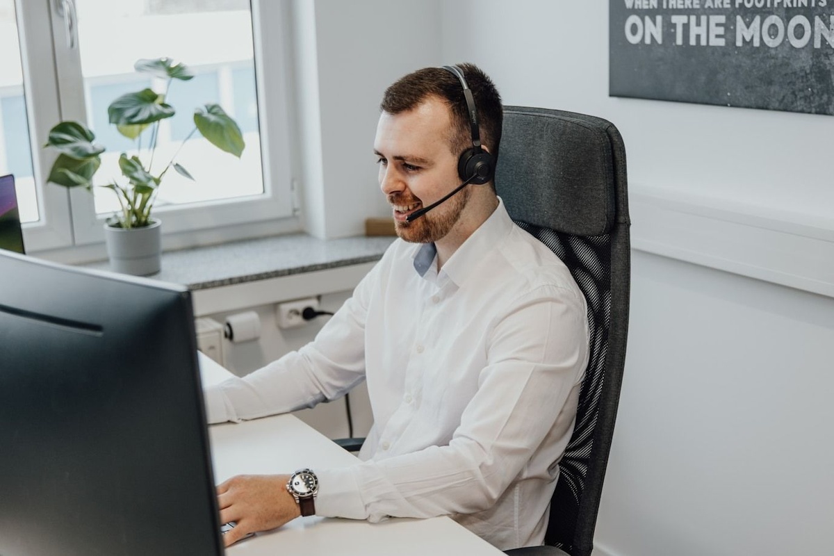 An image of a man in a white shirt with a headset, sat at a desk whilst smiling. 