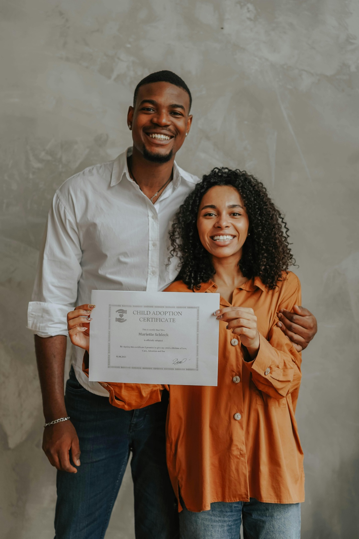 Two people standing indoors and holding up a certificate together, with one person’s arm around the other’s shoulder against a neutral background.