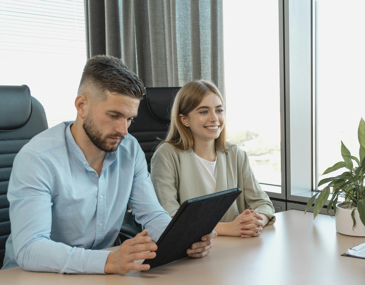 Two people sit at a conference table near large windows, with one person holding a tablet while the other sits with hands folded.