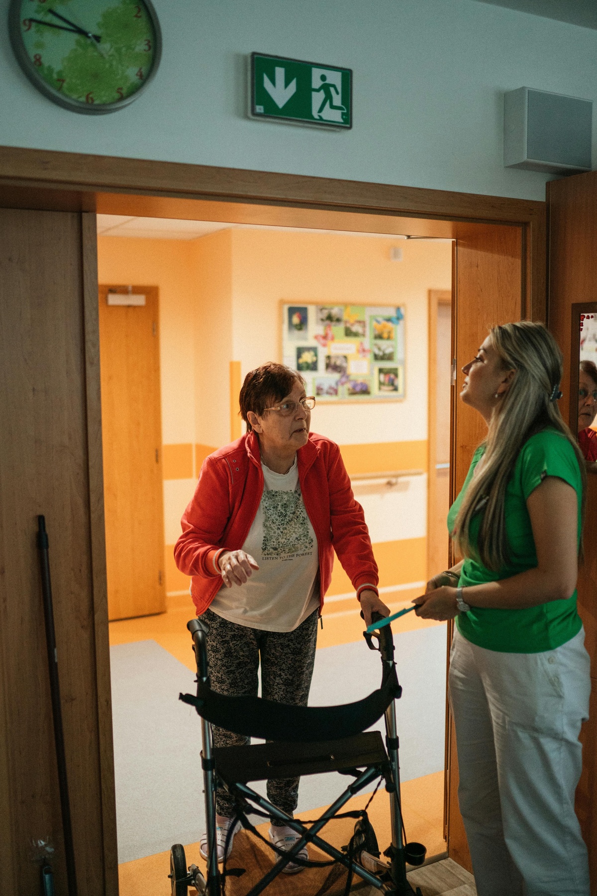 Person using a walker paused in a doorway inside a care or residential setting, speaking with another person standing nearby, with a wheelchair positioned in front and a brightly lit corridor visible beyond the door.