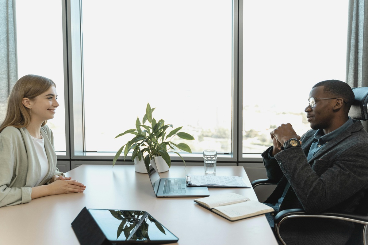 Two people sit across from each other at a desk in a bright office, with a laptop, notebook, glass of water, and a plant placed between them.