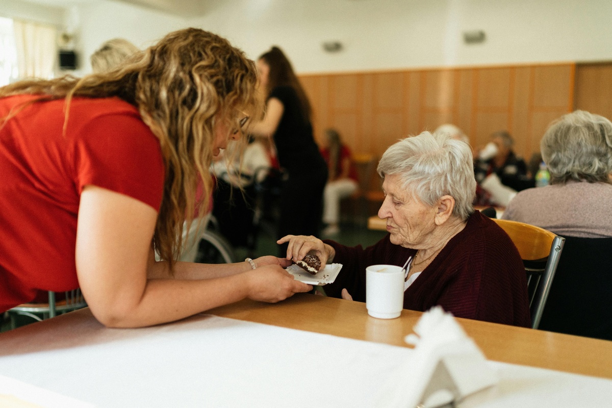 Care worker leaning over a table to assist an older adult seated in a communal dining area, with a mug on the table and other people seated in the background in a care or residential setting.