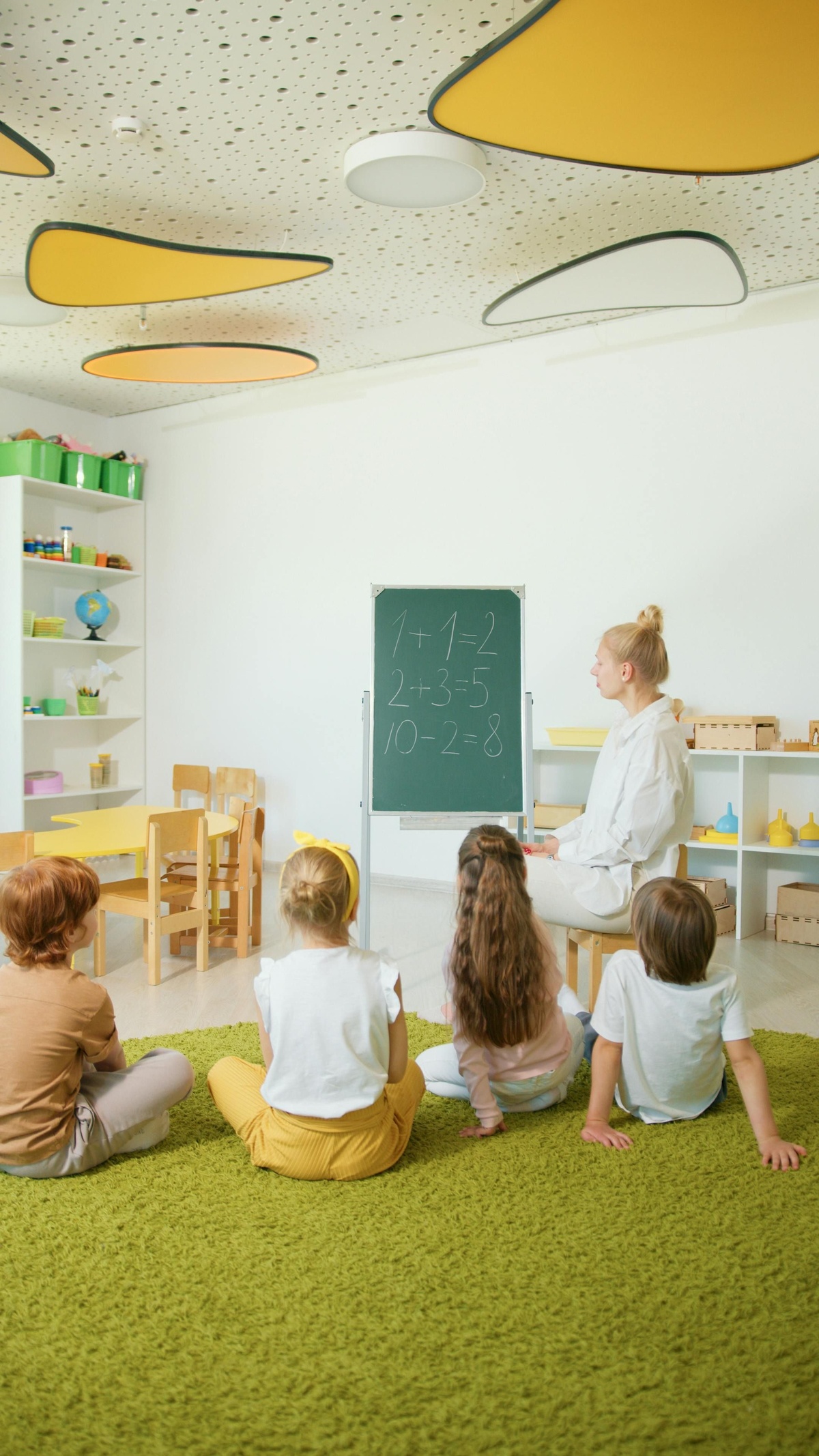 Teacher seated at the front of a bright classroom, using a small chalkboard with maths written on it, while young children sit on the floor listening in a nursery or early‑years learning environment.