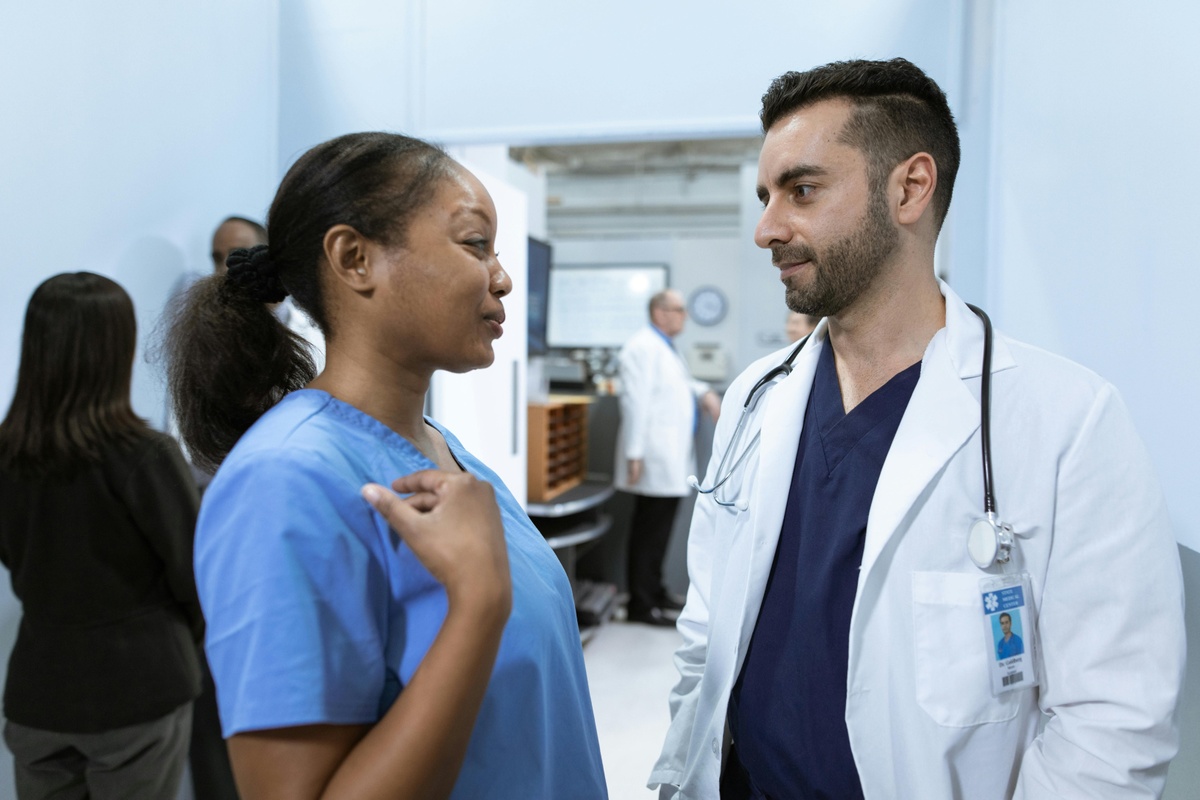 Two healthcare professionals standing in a hospital corridor, wearing scrubs and a lab coat, talking together during a discussion, with other staff and clinical equipment visible in the background.