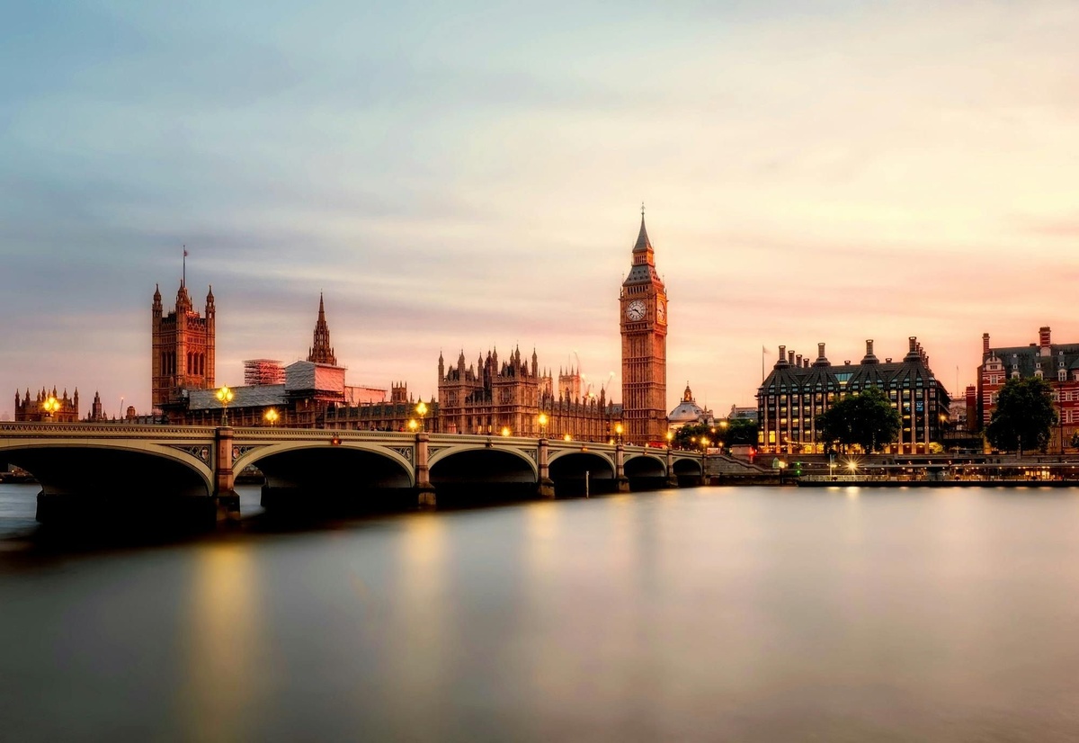 Riverfront view of London at sunset, showing the Palace of Westminster, Big Ben, and Westminster Bridge reflected in the water.