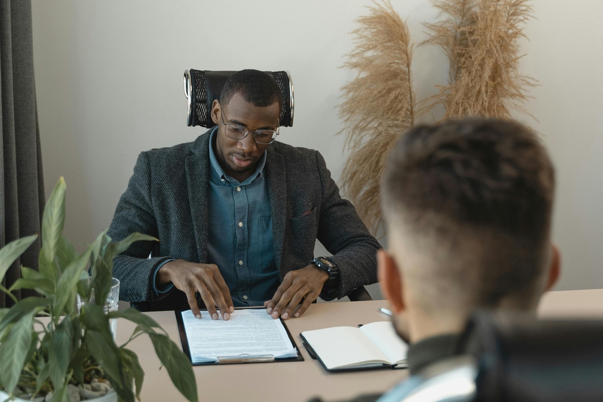 A person in a suit sits at a desk with paperwork, speaking to another person seated across the table, with plants and decor in the background.