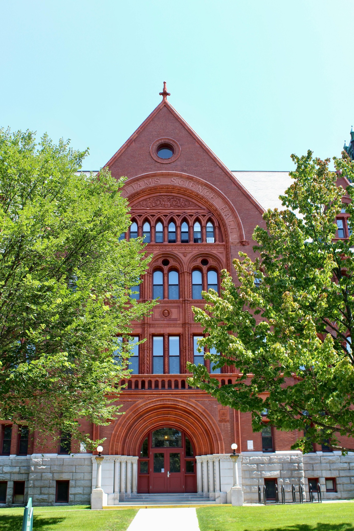 Large red‑brick academic building with arched doorways, decorative stonework, and trees framing the entrance on a sunny day.
