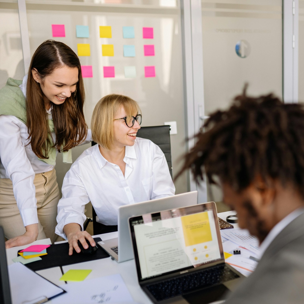 Group of people gathered around a desk in an office, reviewing content on a laptop and printed documents, with colourful sticky notes visible on a glass wall in the background.