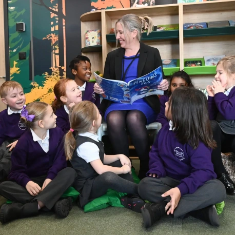 An adult reading a book to a group of primary school pupils seated on cushions in a classroom, with bookshelves and learning resources in the background.