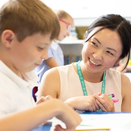 Adult seated beside a pupil at a classroom table, supporting a learning activity using pens and worksheets. Additional pupils and classroom resources are visible in the background, highlighting a collaborative and engaging educational environment.