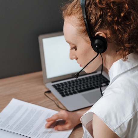Pupil using a headset while reading printed materials at a desk, with a laptop open in the background during a technology-supported learning activity.