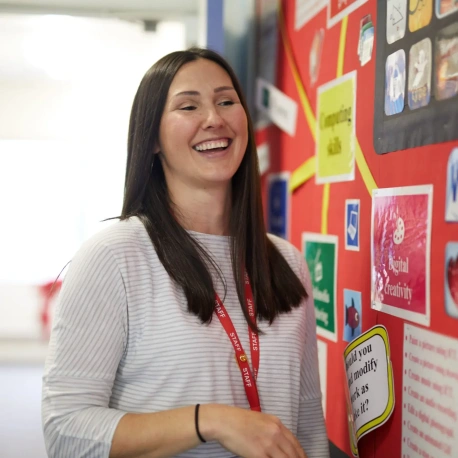 An adult standing beside a classroom display board, with educational posters and learning materials visible in the background.