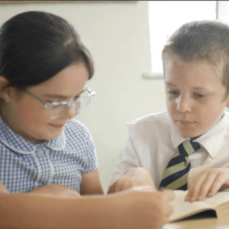 Two pupils reading together at a classroom table, focusing on an open book during a classroom activity.