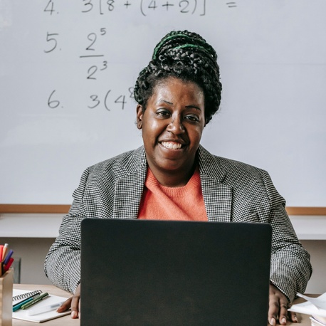 Person seated behind a laptop in a classroom, with a whiteboard showing maths equations in the background.