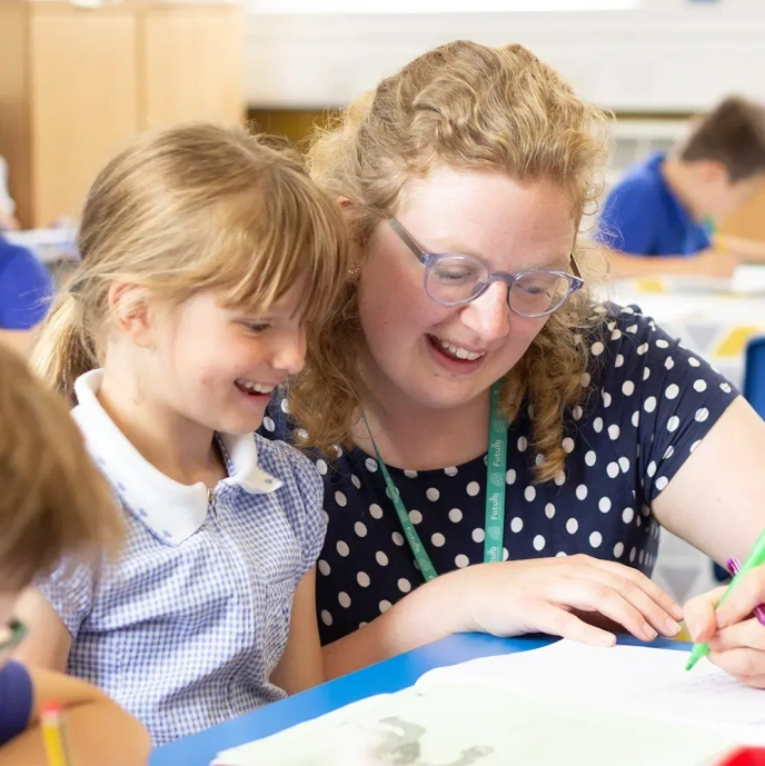 Adult seated beside a pupil at a classroom table, working together on a written activity. Paper, pens and learning materials are spread across the table, while other pupils and classroom resources are visible in the background, highlighting a supportive and collaborative learning environment.