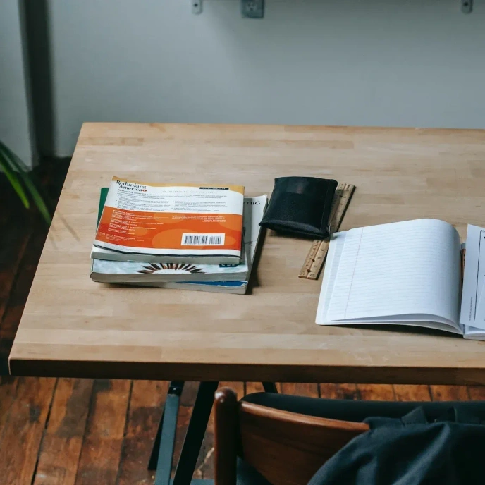 Books, a notebook, a ruler and a wallet arranged on a wooden desk in a classroom or study setting.