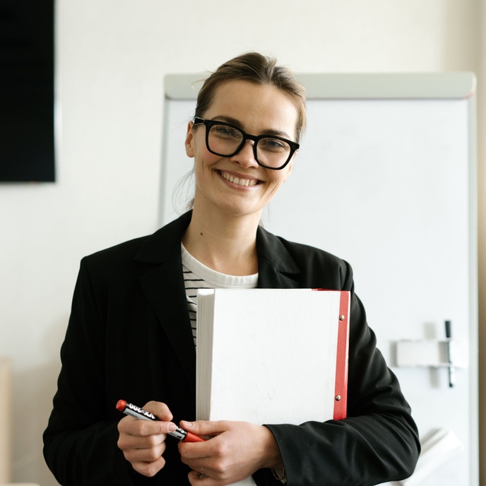 Person holding a folder and marker while standing in a classroom, facing the camera with a whiteboard visible in the background.