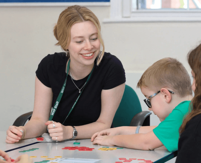 Adult supporting a child with a learning activity at a table while other pupils take part nearby.