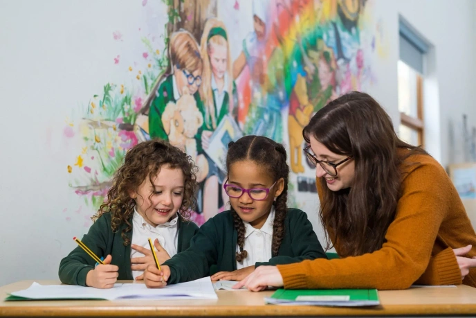 Adult supporting two children as they draw and write together at a table in a classroom with a colourful mural in the background.