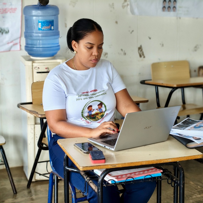 Person seated at a classroom desk, working on a laptop with a phone and notebooks nearby.