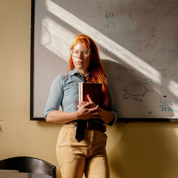 Person standing in front of a classroom whiteboard holding a book, with educational notes and diagrams visible in the background.