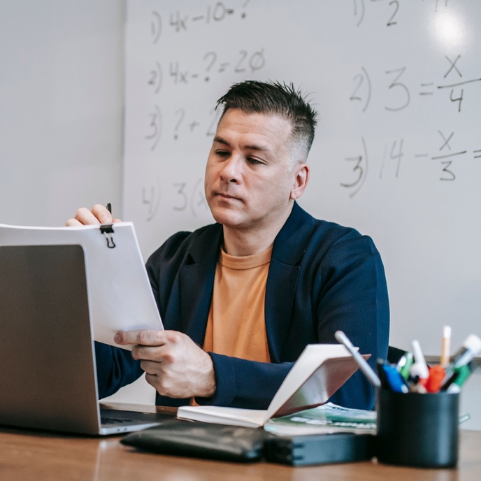 Person seated at a desk reviewing a document beside a laptop, with a whiteboard and office supplies in the background.